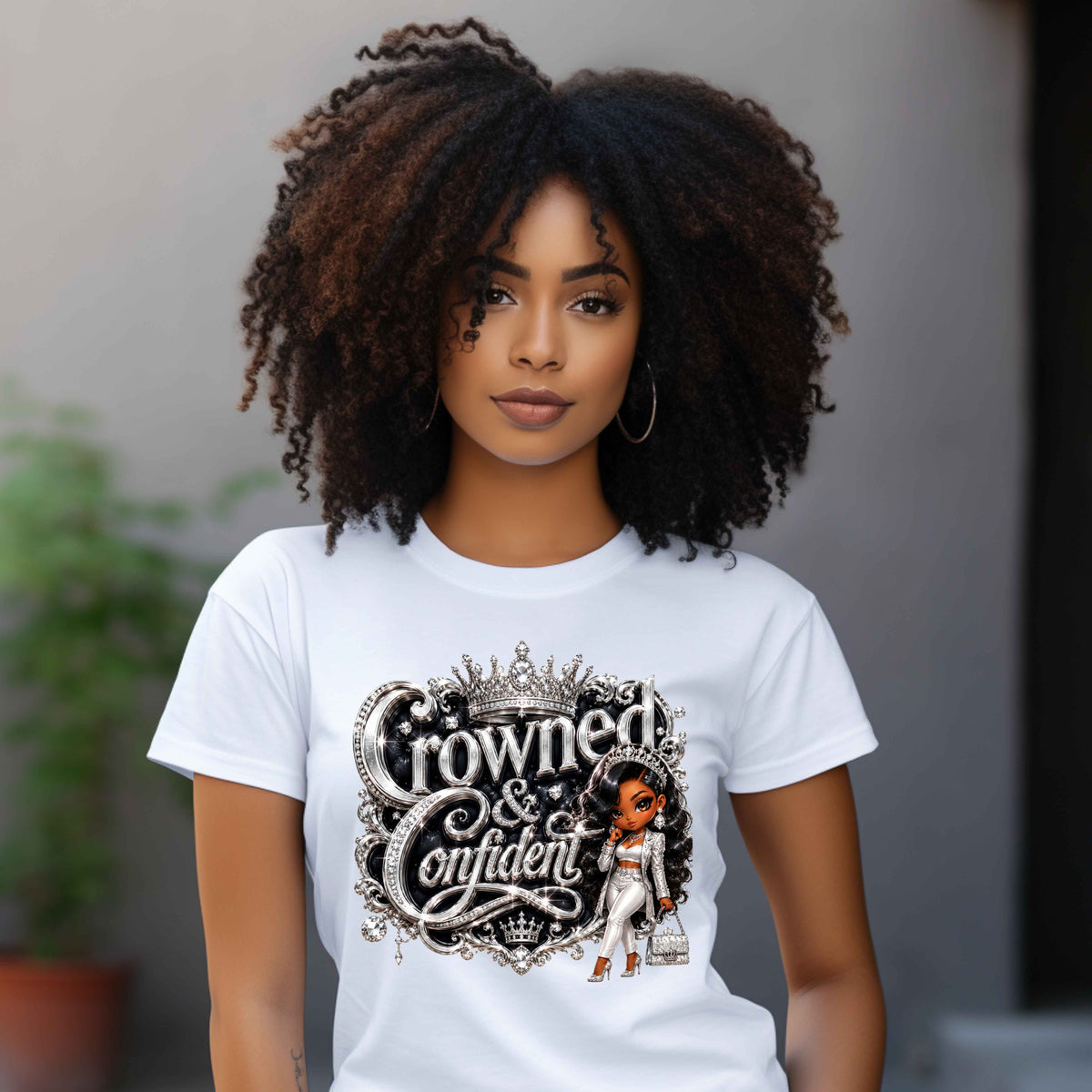 A woman with voluminous dark curly hair wearing a white graphic t-shirt with 'Crowned & Confident' printed in an ornate black and silver font, featuring a small illustration of a woman in a white gown. The background is a soft-focus gray wall with a hint of greenery.