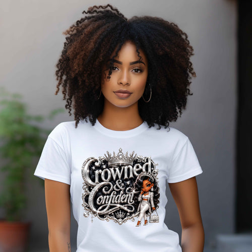 A woman with voluminous dark curly hair wearing a white graphic t-shirt with 'Crowned & Confident' printed in an ornate black and silver font, featuring a small illustration of a woman in a white gown. The background is a soft-focus gray wall with a hint of greenery.