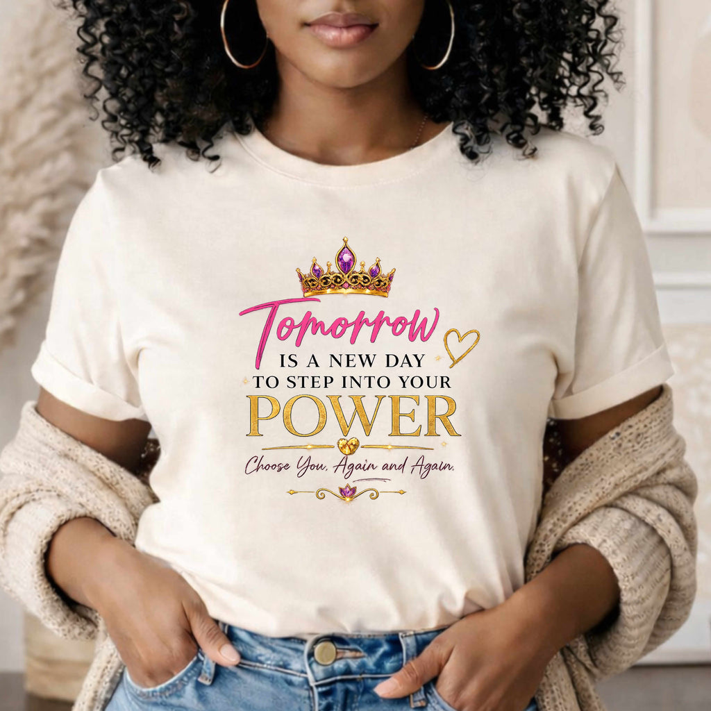 A woman wearing a beige graphic t-shirt with a motivational quote printed in pink and gold text under a gold crown graphic. The text reads 'Tomorrow IS A NEW DAY TO STEP INTO YOUR POWER Choose You, Again and Again.

Details