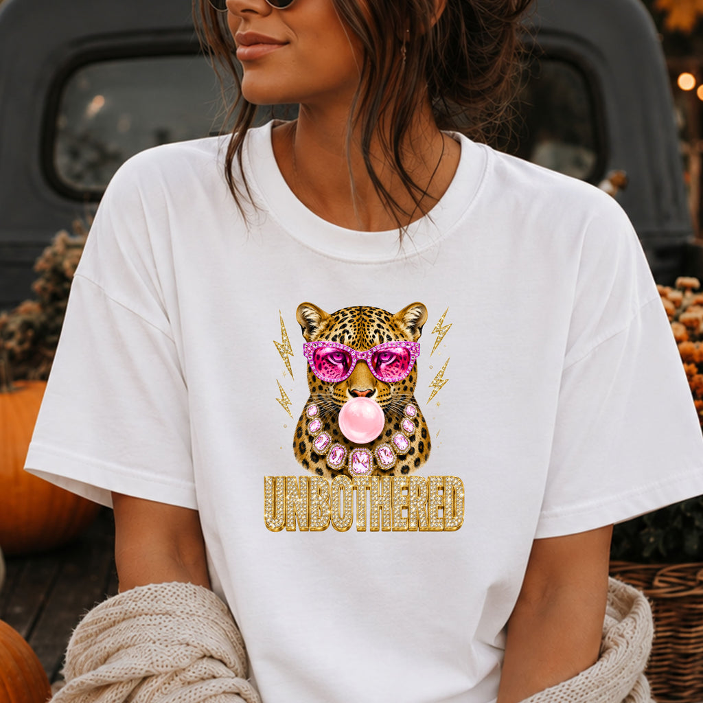 A woman wearing a white graphic t-shirt featuring a leopard wearing pink sunglasses and blowing pink bubblegum, with the word 'UNBOtherED' printed below in gold glitter-style lettering.